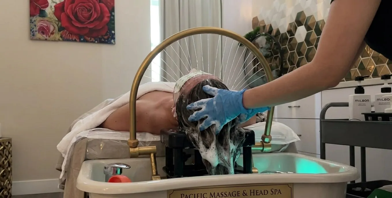 Therapist working a guest's scalp at the reclining hydrotherapy basin under the overhead rinse fixture during a head spa session