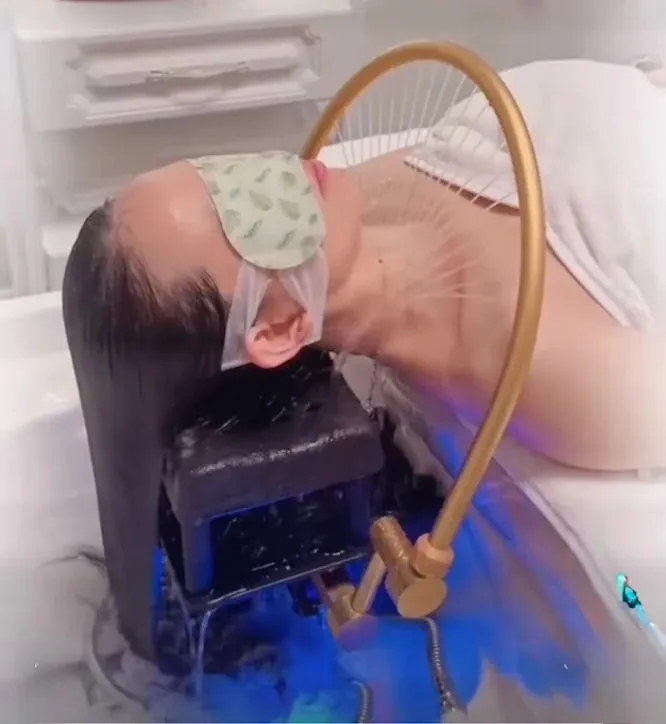 Warm water running from the overhead arch fixture through a guest's hair during the rinse stage of a head spa session